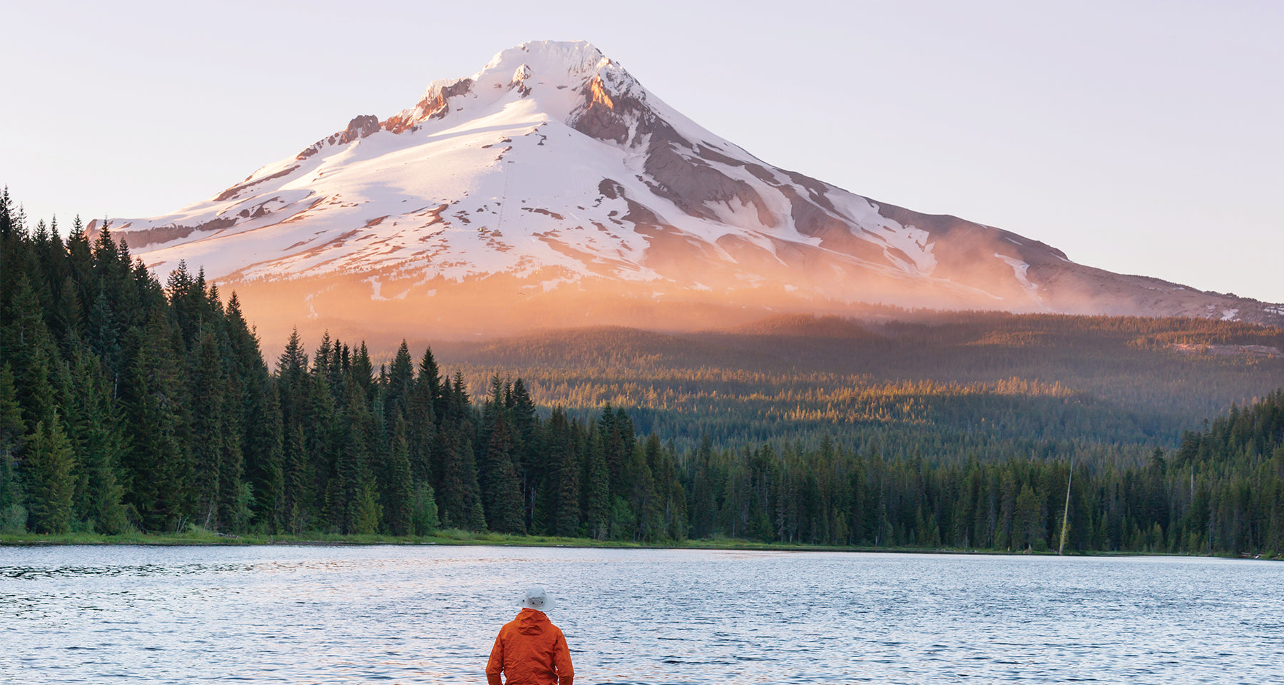 Snow-capped mountain in the distance framed by a stand of evergreen trees before a calm blue lake. A man wearing a white boating hat and orange rain slicker stands in the foreground with his back to the viewer.
