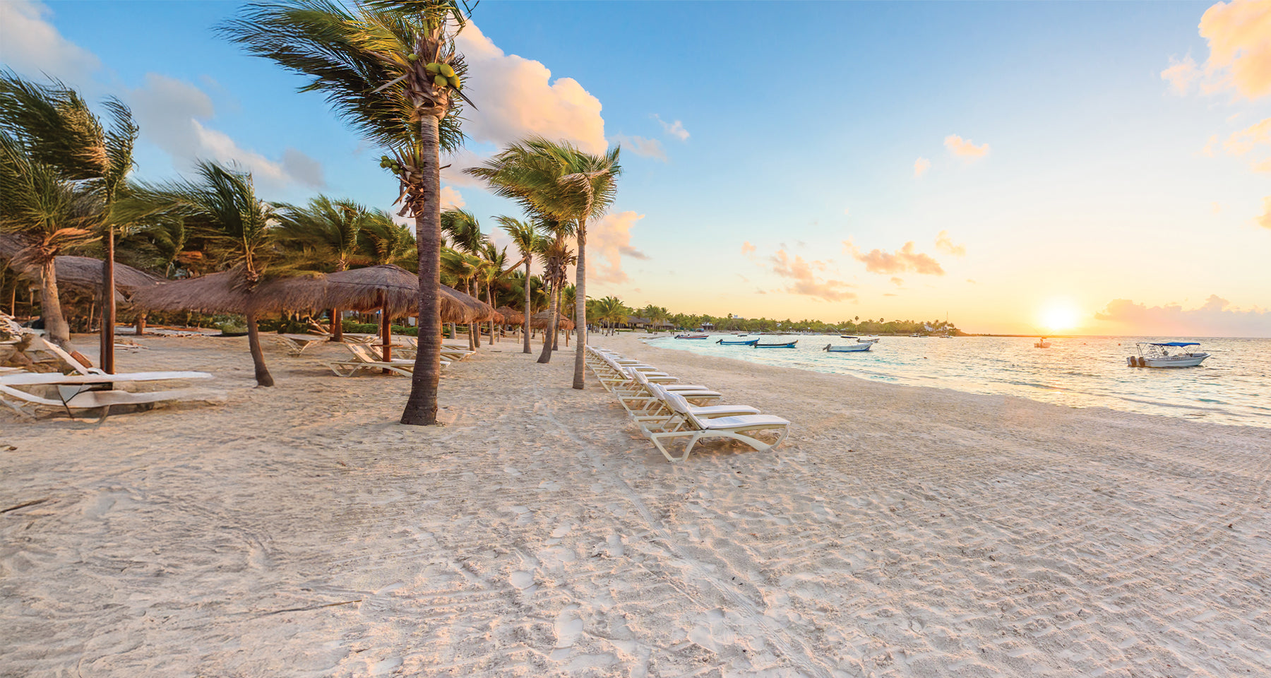Beach scene with palm trees, lounge chairs, and boats at sunset.